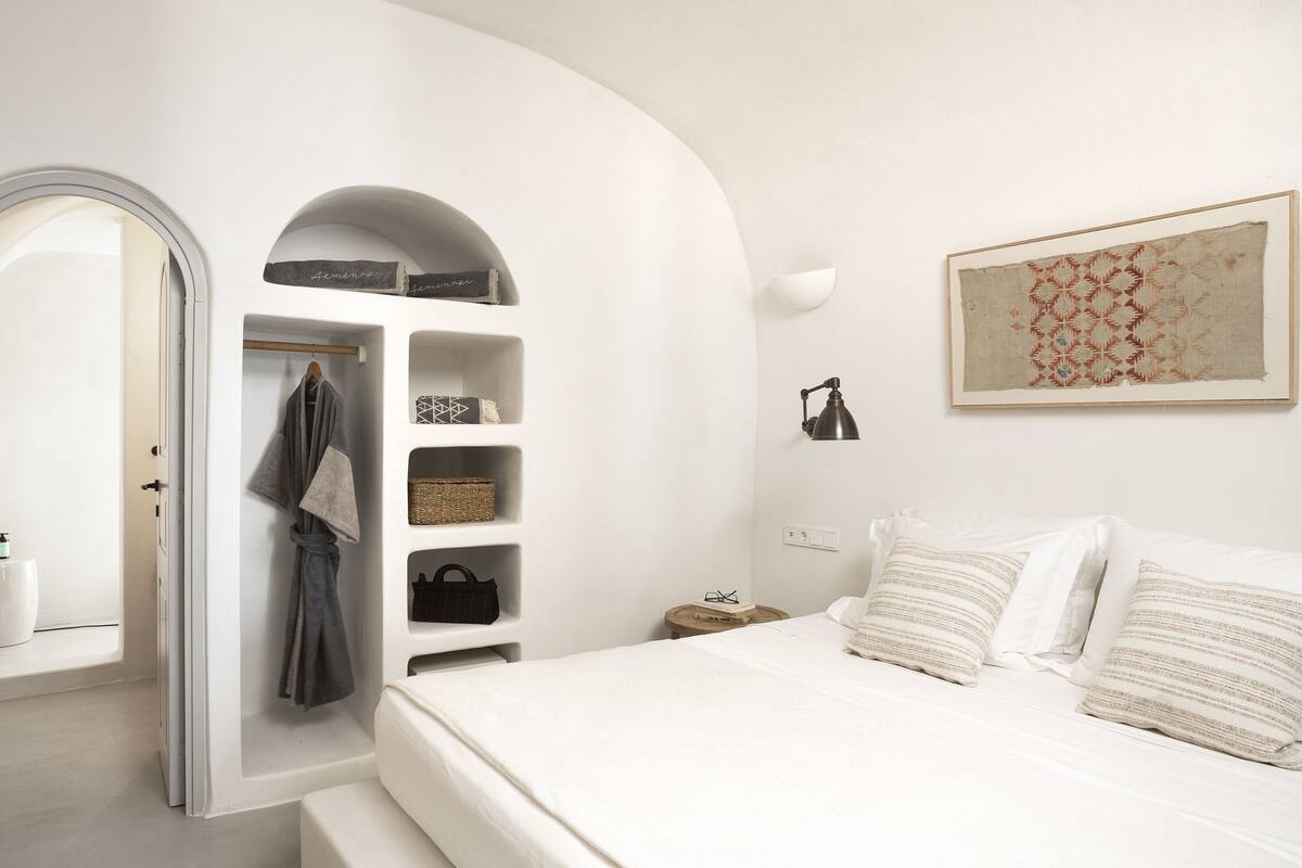 Un dormitorio minimalista con cama blanca, una bata colgada en un perchero y una estantería con cajas de almacenamiento y una cesta.