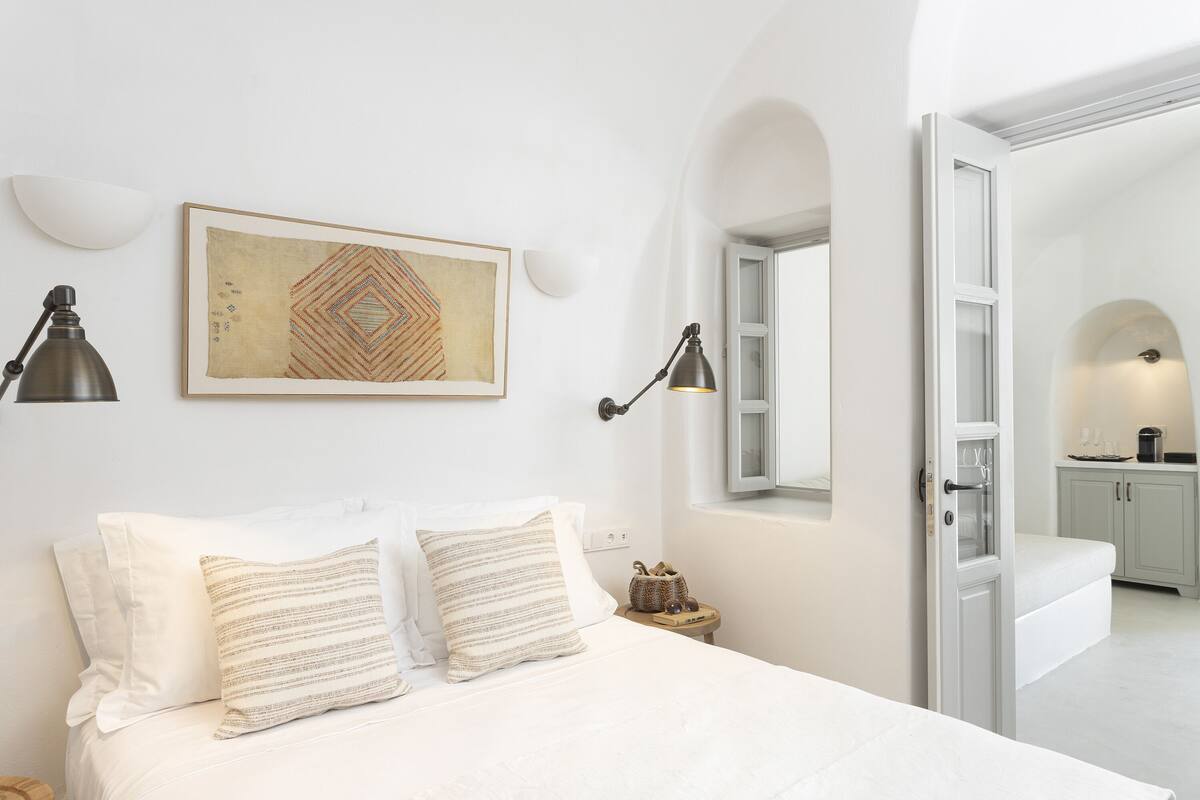 Un dormitorio con cama, dos almohadas, una mesita de noche, lámpara, un cuadro en la pared y una puerta abierta que da al baño.