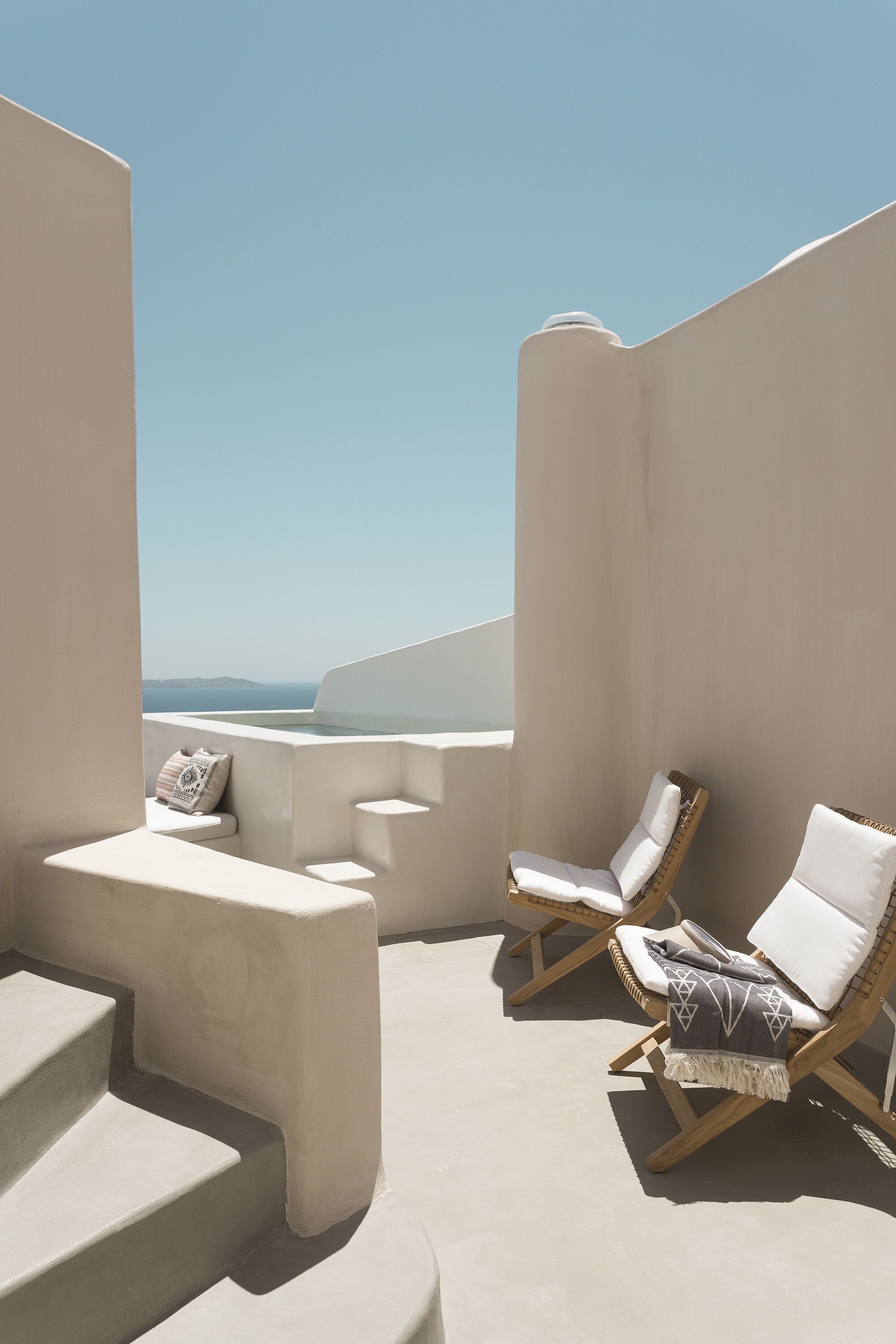 Une terrasse ensoleillée aux murs blancs, offrant une vue sur la mer. On y trouve deux chaises longues et une petite table avec un coussin.