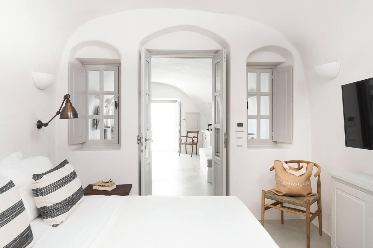 Un dormitorio con cama, lámpara de noche, silla con cojín y televisión en la pared.