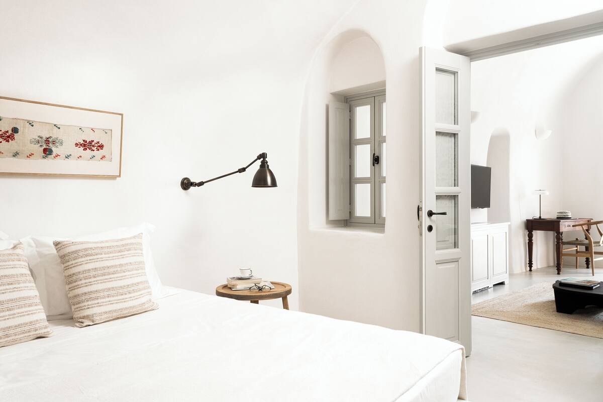 Un dormitorio con cama, almohadas, mesita de noche con lámpara, una obra de arte enmarcada y una puerta que da a otra habitación.