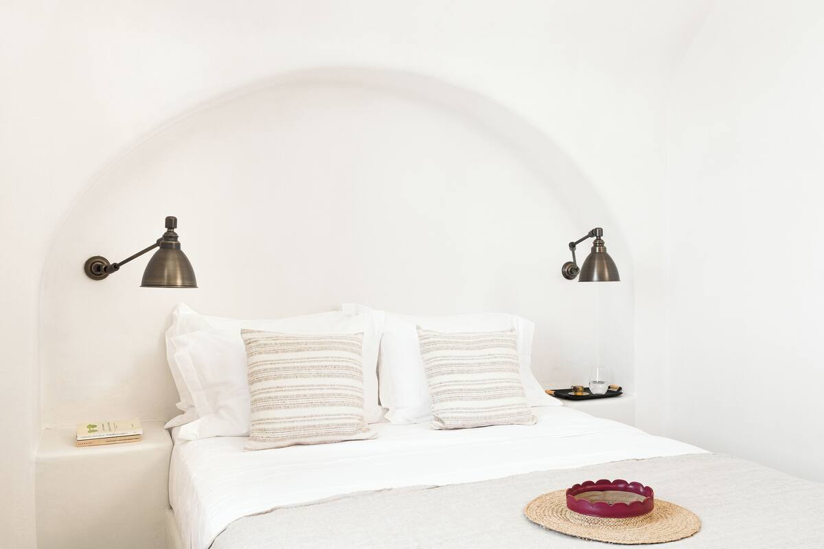 Una cama bien tendida con sábanas blancas y almohadas a rayas, dos lámparas fijadas a la pared y una mesita de noche con un libro y un vaso.