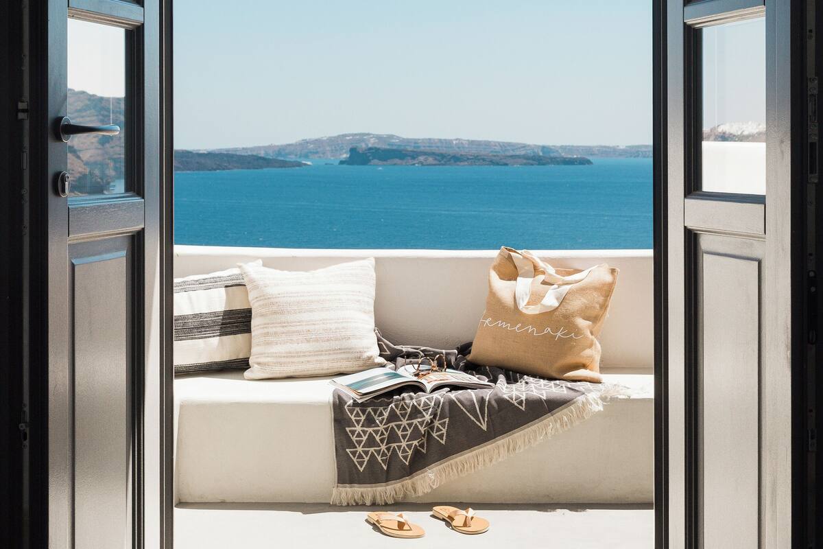 Una puerta abierta que da a un balcón con vistas al mar y colinas al horizonte.