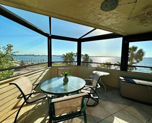 A sunlit patio with a round glass table, wicker chairs, and a view of the sea. - Bayview Condo D - Anna Maria Island - Condo 2 Bedroom/ 2 Bath bay view, maximum occupancy of 4 people.  Community pool. Distance to the Beach: 1 block (Bradenton Beach)