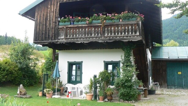 Une maison en bois à deux étages, dotée d’un balcon et entourée de verdure et de plantes en pot.