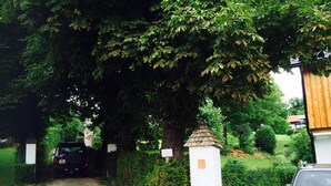 Une allée privée fermée par un portail, avec une voiture garée à l’intérieur, entourée d’une végétation luxuriante et d’un bâtiment blanc surmonté d’une petite structure ressemblant à une tour.