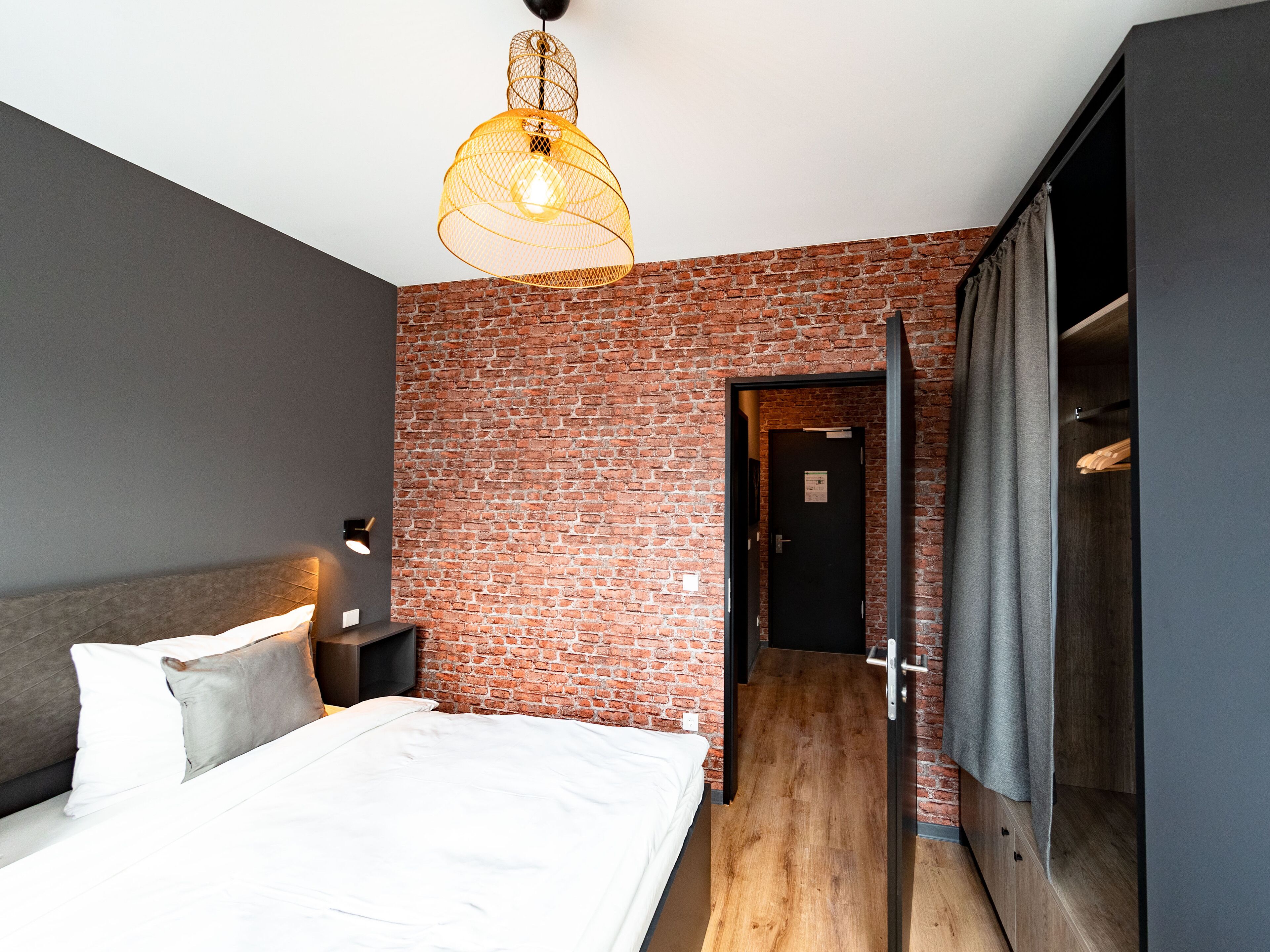 Un dormitorio moderno con pared de ladrillo, cama con ropa de cama blanca, lámpara colgante y suelo de madera.