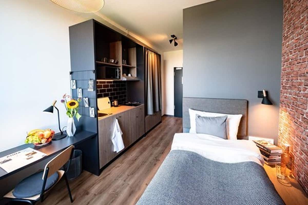 Quarto de hotel compacto com cozinha compacta, cama com cabeceira cinza, escrivaninha com cadeira e mesa de jantar com uma fruteira.