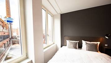 Una habitación de hotel moderna con una cama grande, una pared de acento oscura y una ventana con vistas a un paisaje urbano.