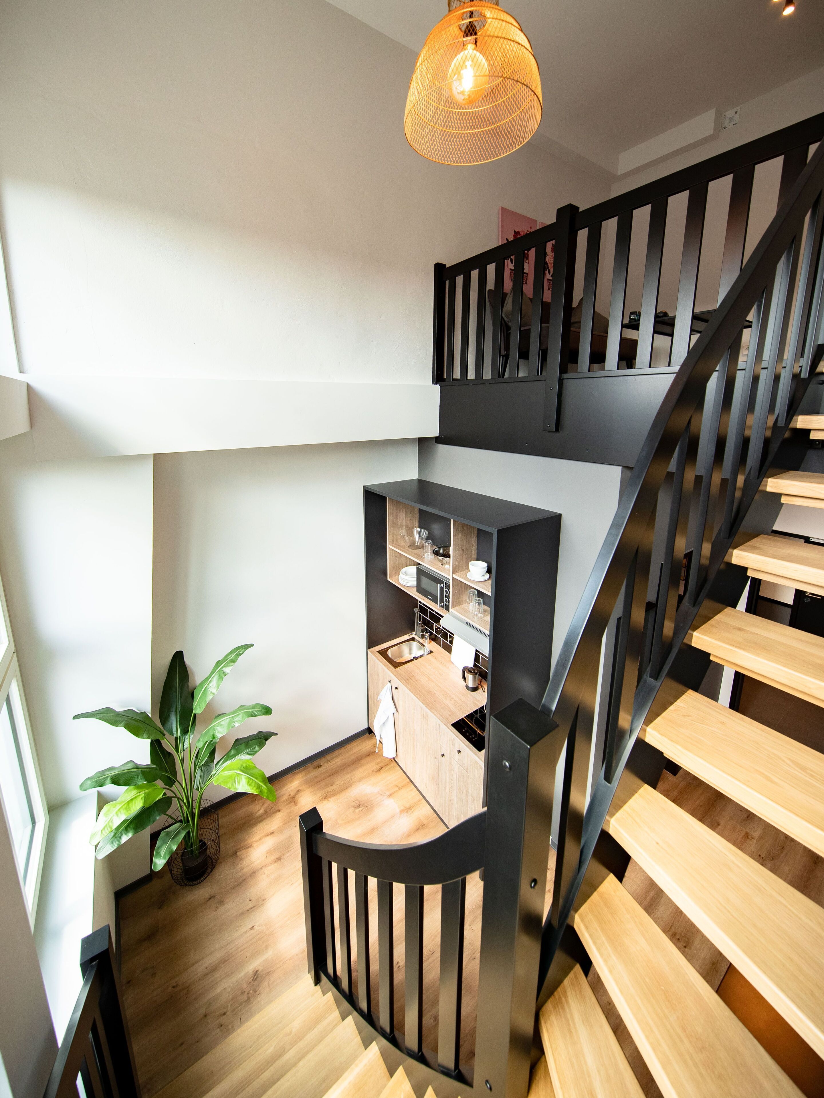 A modern kitchen with wooden flooring, a black staircase, and a hanging light fixture.