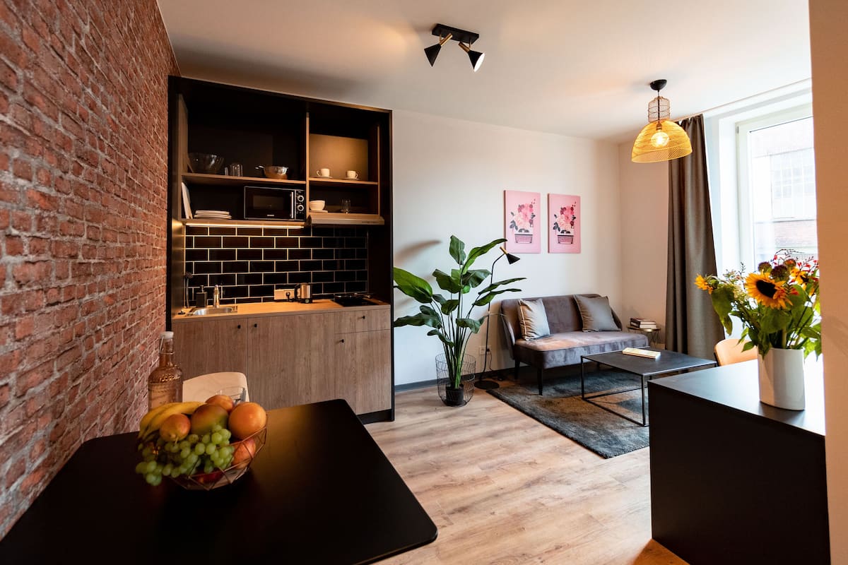 Cozinha moderna com parede de tijolos, armários de madeira e painel de azulejos pretos. Há uma mesa de jantar com um cesto de frutas, um sofá e uma mesa de centro na sala de estar.