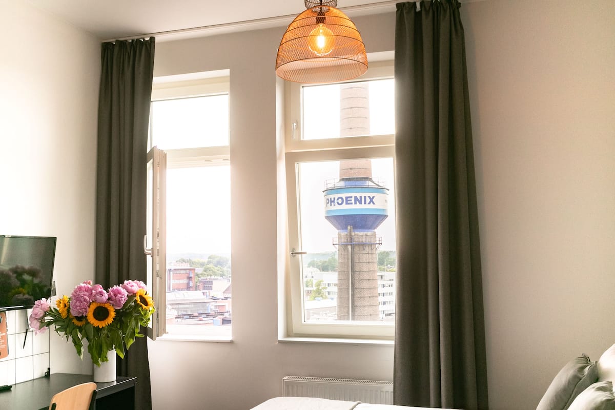 Quarto de hotel com cama, escrivaninha com computador, cadeira, um vaso de flores e janela com vista para um tanque d'água identificado como “PHOENIX”.