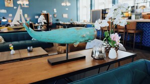 Una escultura de ballena color turquesa sobre una mesa, orquídeas blancas en un florero y una silla azul en un restaurante.