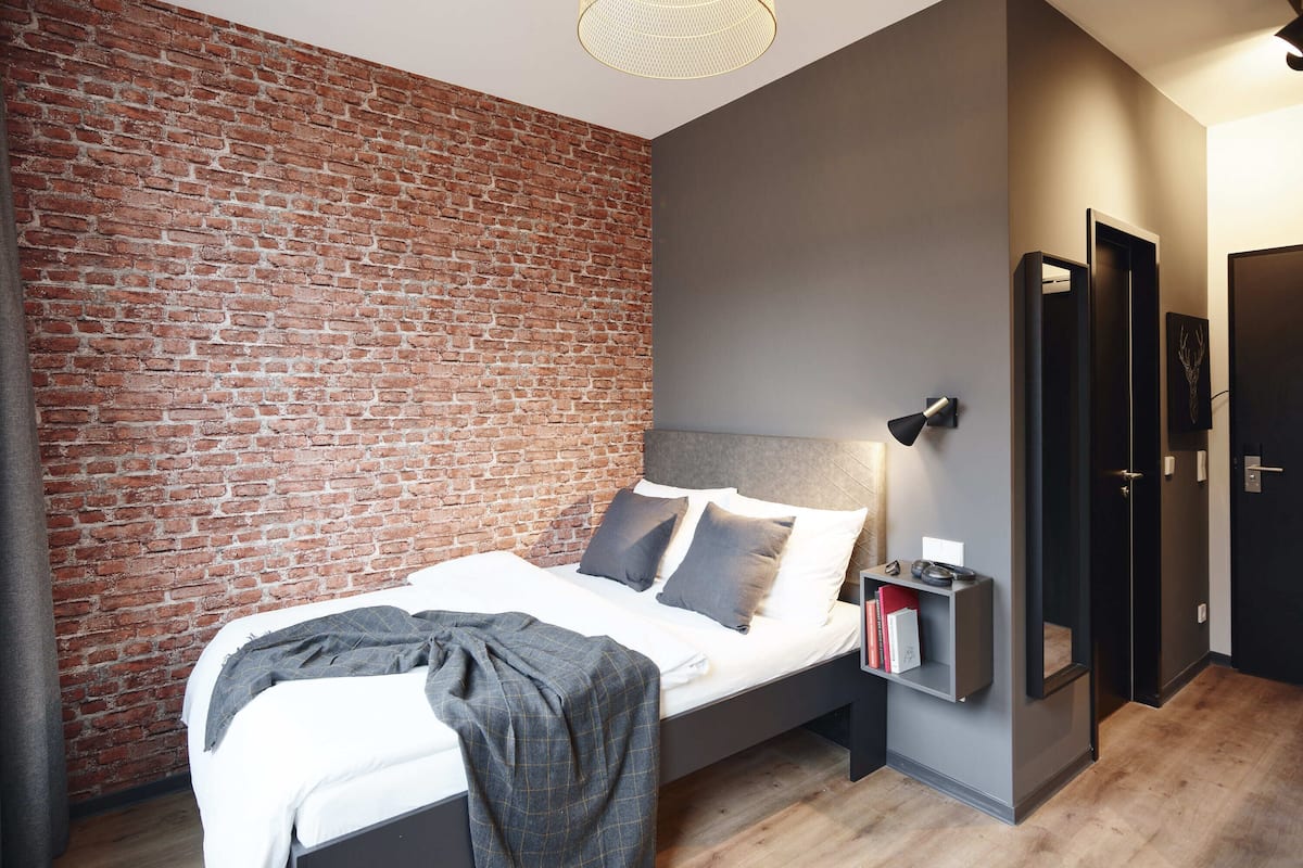 Quarto moderno com parede de tijolos, uma cama com almofadas, mesa de cabeceira com livros e uma porta.
