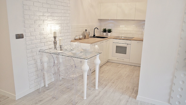 A modern kitchen with white brick walls, a wooden countertop, and a glass dining table with clear chairs. - Warsaw Concierge Opera Apartment (Warsaw)