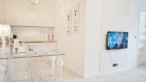 A modern kitchen with white cabinetry, a glass dining table with clear chairs, and a mounted TV on the wall. - Warsaw Concierge Opera Apartment (Warsaw)