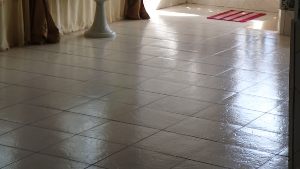 A room with a tiled floor, a potted plant, a window with curtains, and a decorative wall. - Al Djadid (Domoni)