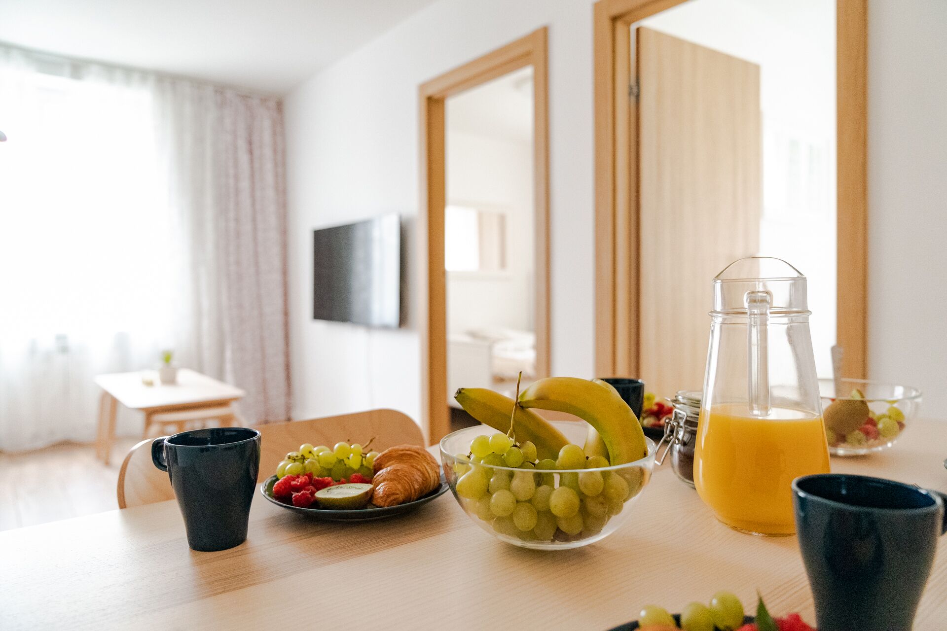 Una mesa de madera con un bol de fruta, cruasanes y una jarra de jugo.