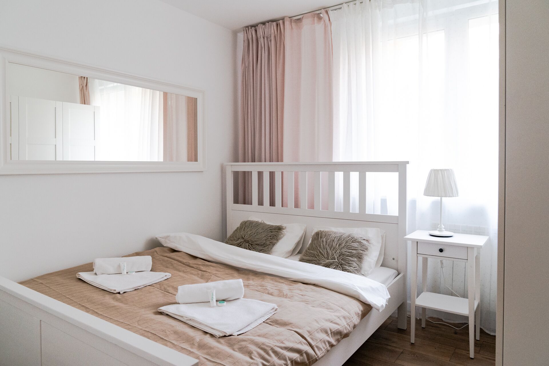 Een net opgemaakt bed met witte lakens en een beige sprei, een nachtkastje met een lamp en een raam met dunne gordijnen.