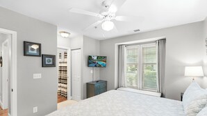Ein Schlafzimmer mit Bett, Deckenventilator, Fernseher, Fenster mit Jalousien und Nachttisch.