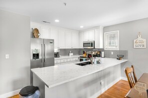 A modern kitchen with white cabinets, stainless steel appliances, a marble countertop, and a wooden dining table.