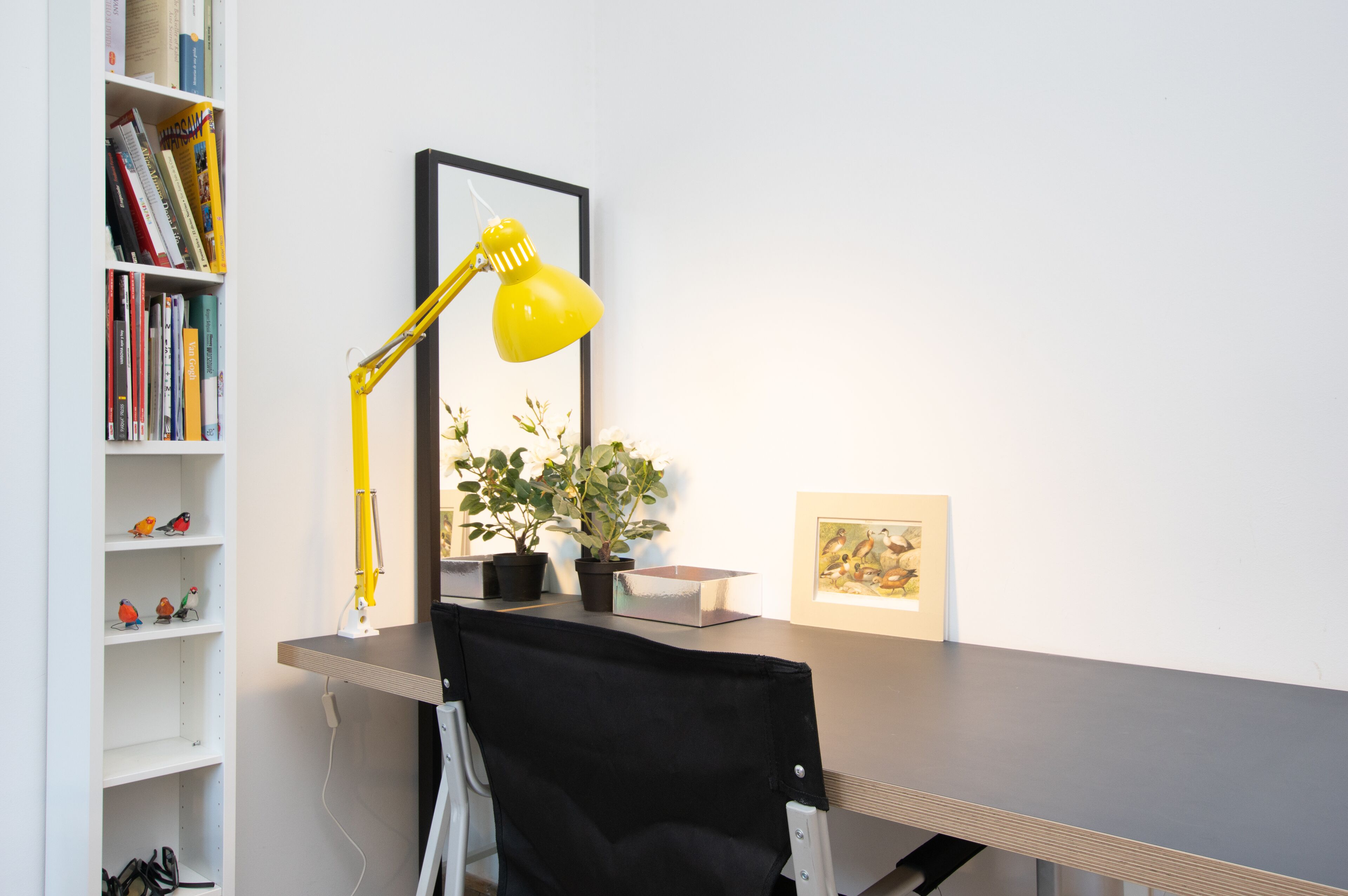 A desk with a yellow desk lamp, a potted plant, and a framed picture. There is a bookshelf with books and decorative items.