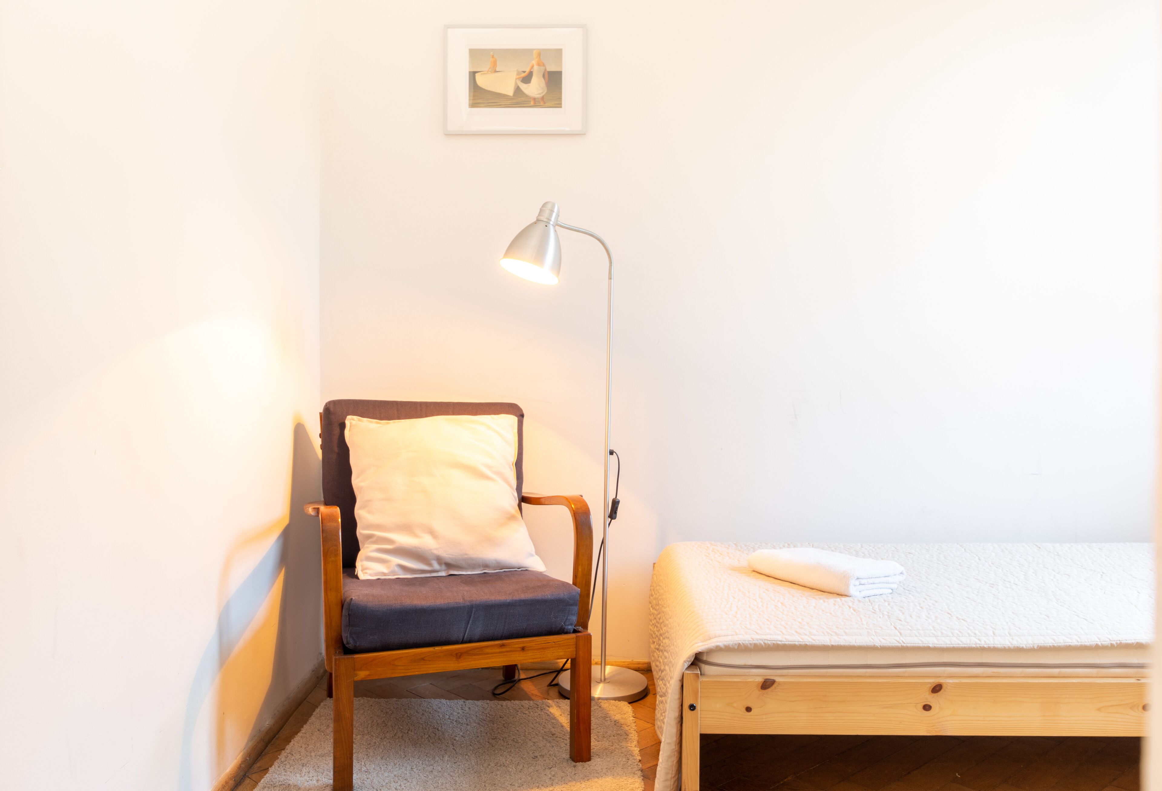 A small, neatly arranged room with a single bed, a chair with a cushion, a floor lamp, and a framed picture on the wall.