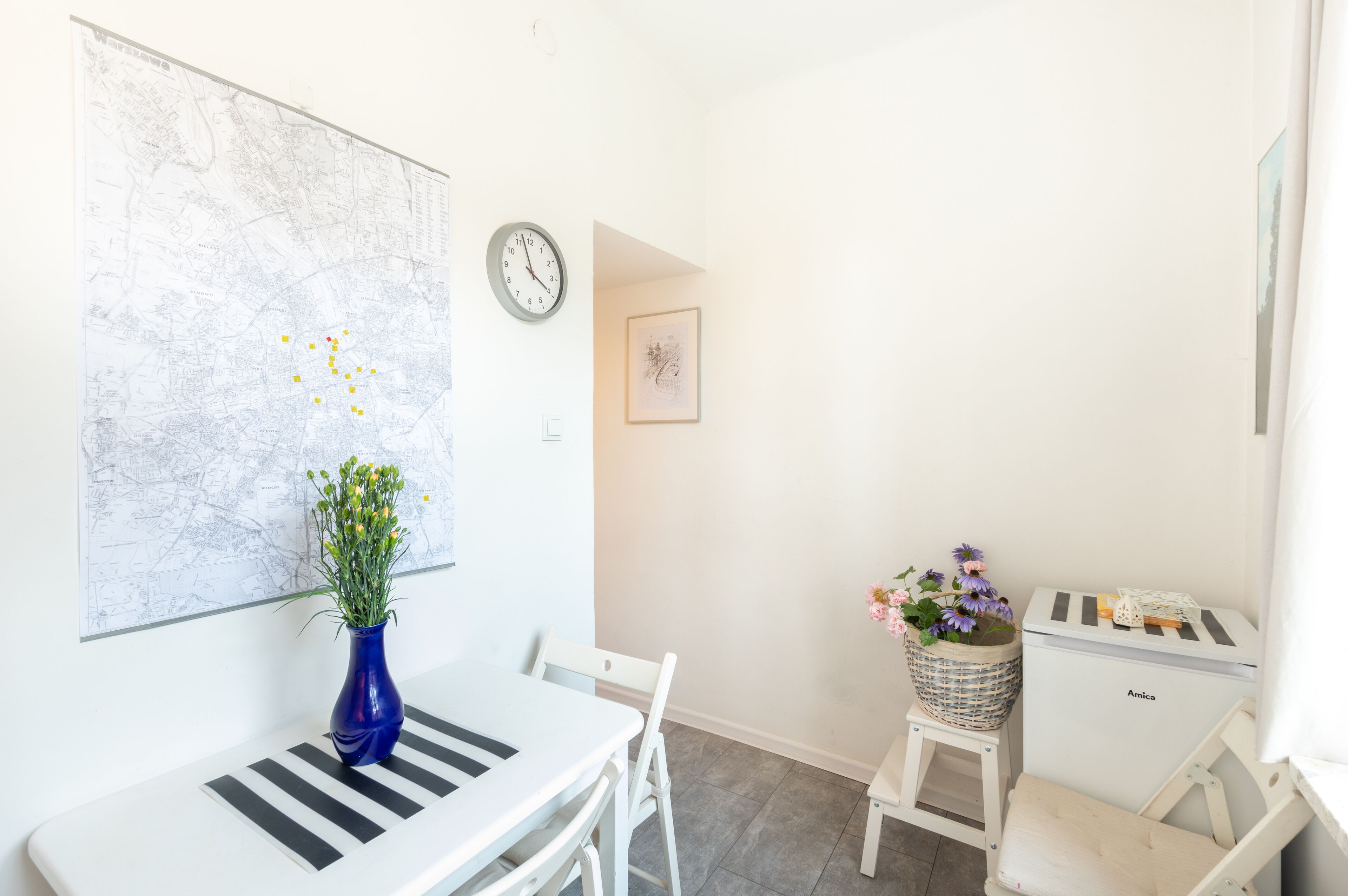 Ein Zimmer mit einem weißen Tisch, einer blauen Vase mit Blumen, einer Karte an der Wand, einer Uhr und einem kleinen Hocker mit einer Blumenkorb.