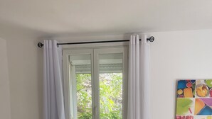 Ein Zimmer mit Fenster, Heizkörper, einem grauen Sofa mit floralen Kissen, einem kleinen Tisch mit Stuhl und einem farbenfrohen Kunstwerk an der Wand.