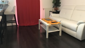 A living room with a white sofa, a red curtain, a small table with a laptop and remote controls, a potted plant, and a radiator. - Disneyland paris esprit (Bussy-Saint-Georges)