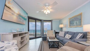 A spacious living area with a ceiling fan, a large painting, a sofa, a coffee table, and a view of the ocean through large windows.