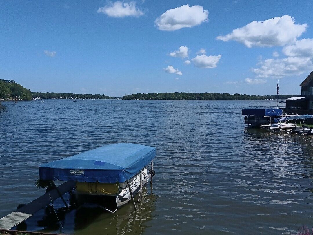 Un barco amarrado con toldo azul, una casa pequeña con una bandera y agua tranquila bajo un cielo despejado.