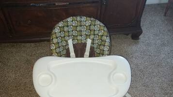 A white baby rocker with a patterned chair behind it, placed on a carpeted floor.