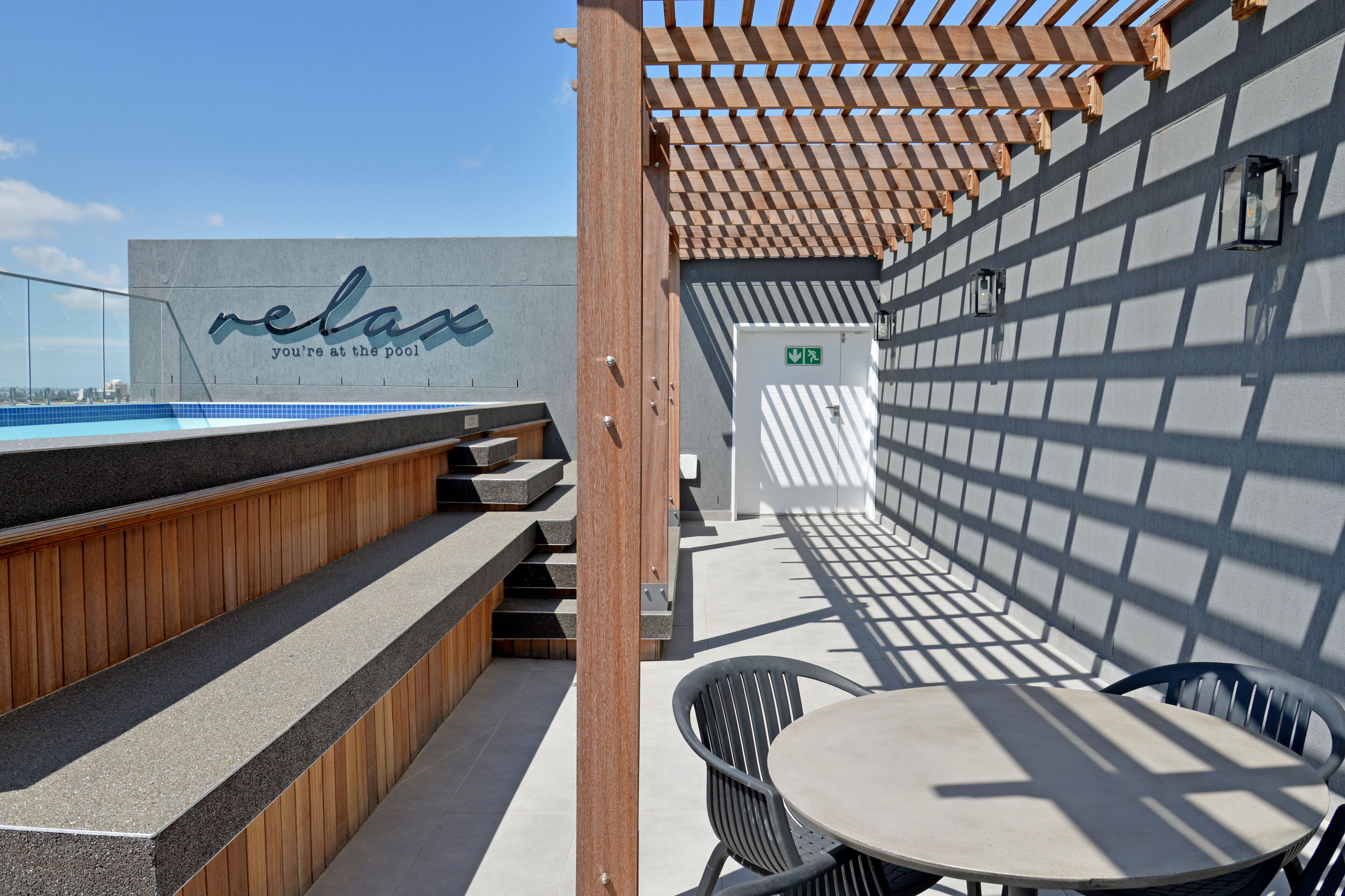 A rooftop pool area with a wooden pergola, a pool, and a seating area with a table and chairs.