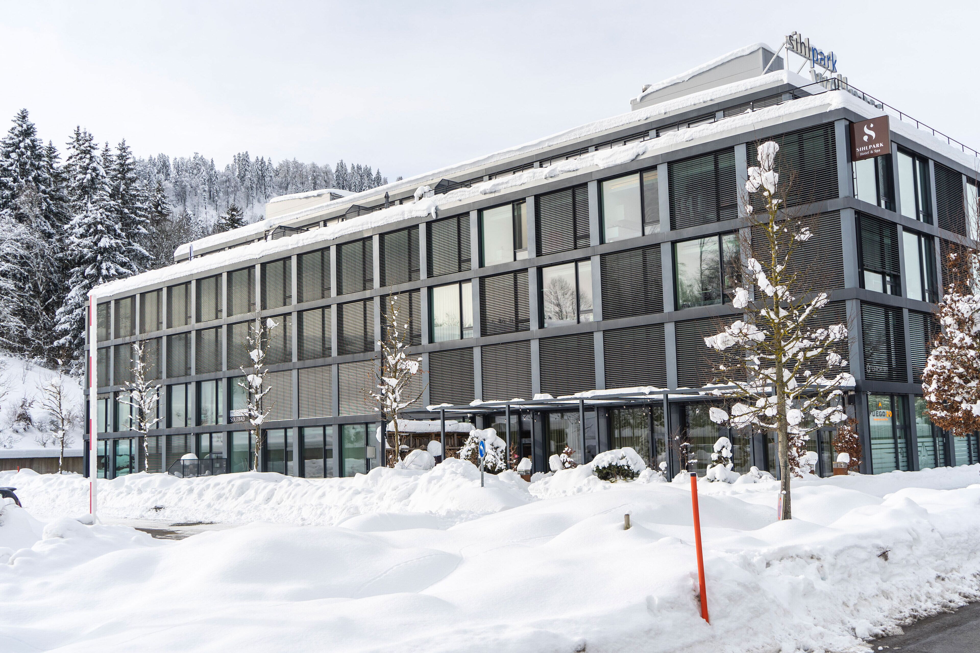 Un edificio moderno con ampie finestre e tetto piano, circondato da terreno e alberi innevati.