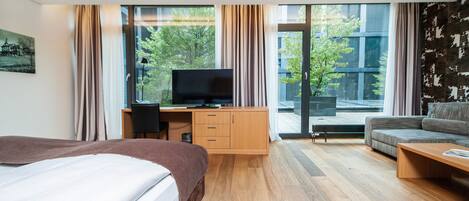 Une chambre d’hôtel moderne dotée d’un grand lit, d’un bureau avec une télévision, d’un canapé et d’une grande fenêtre donnant sur des arbres et des immeubles.