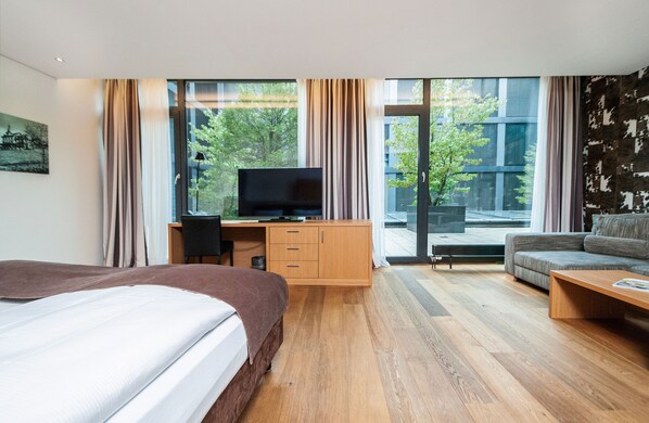 Habitación de hotel moderna con una cama grande, un escritorio con televisor, un sofá y un ventanal que ofrece vistas a árboles y edificios.