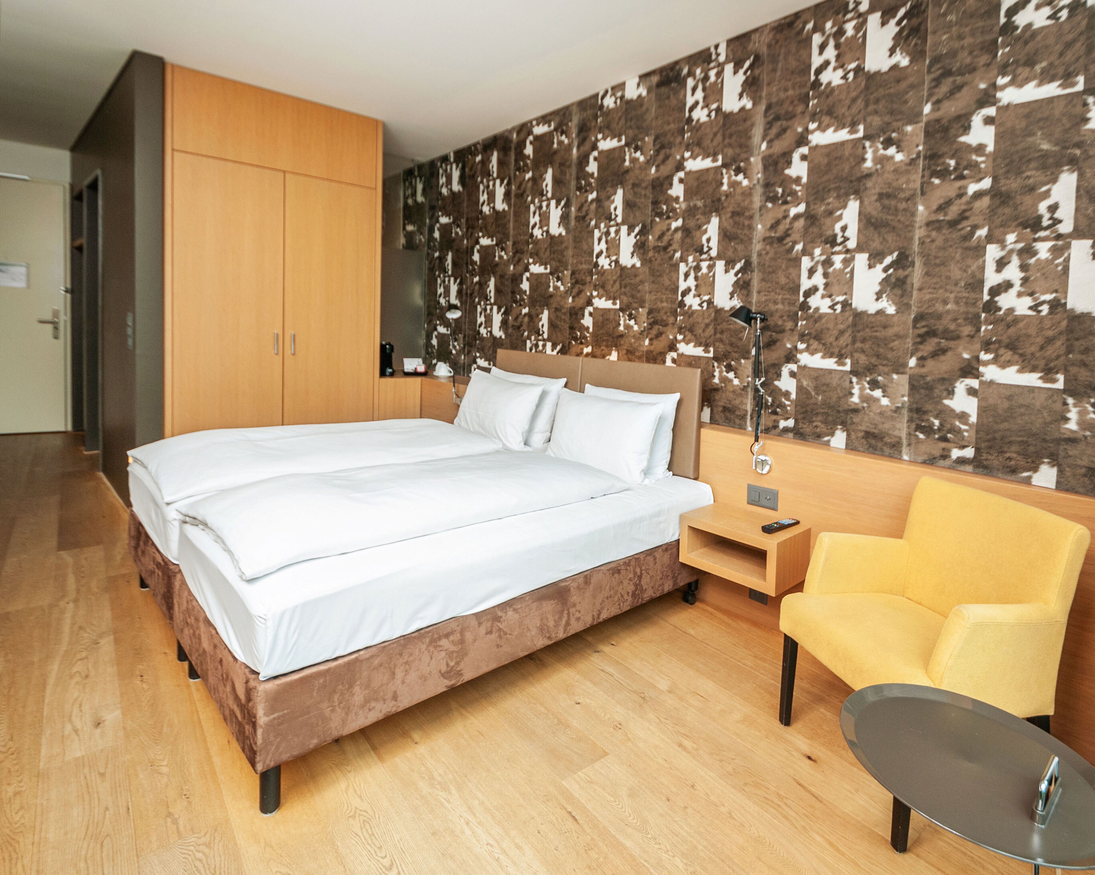 A modern hotel room with a large bed, wooden wardrobe, a desk with a lamp, and a yellow armchair.