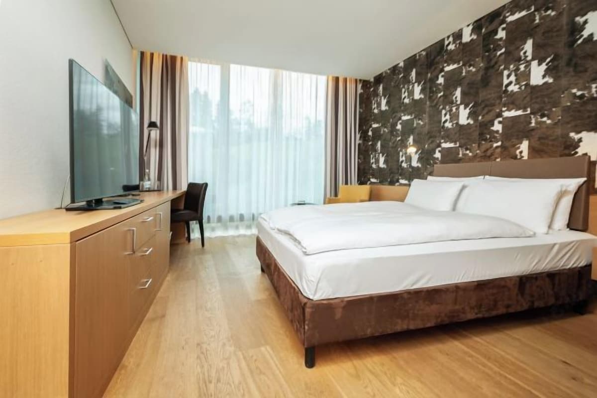 Una habitación de hotel con cama, escritorio con televisor y un ventanal con cortinas.