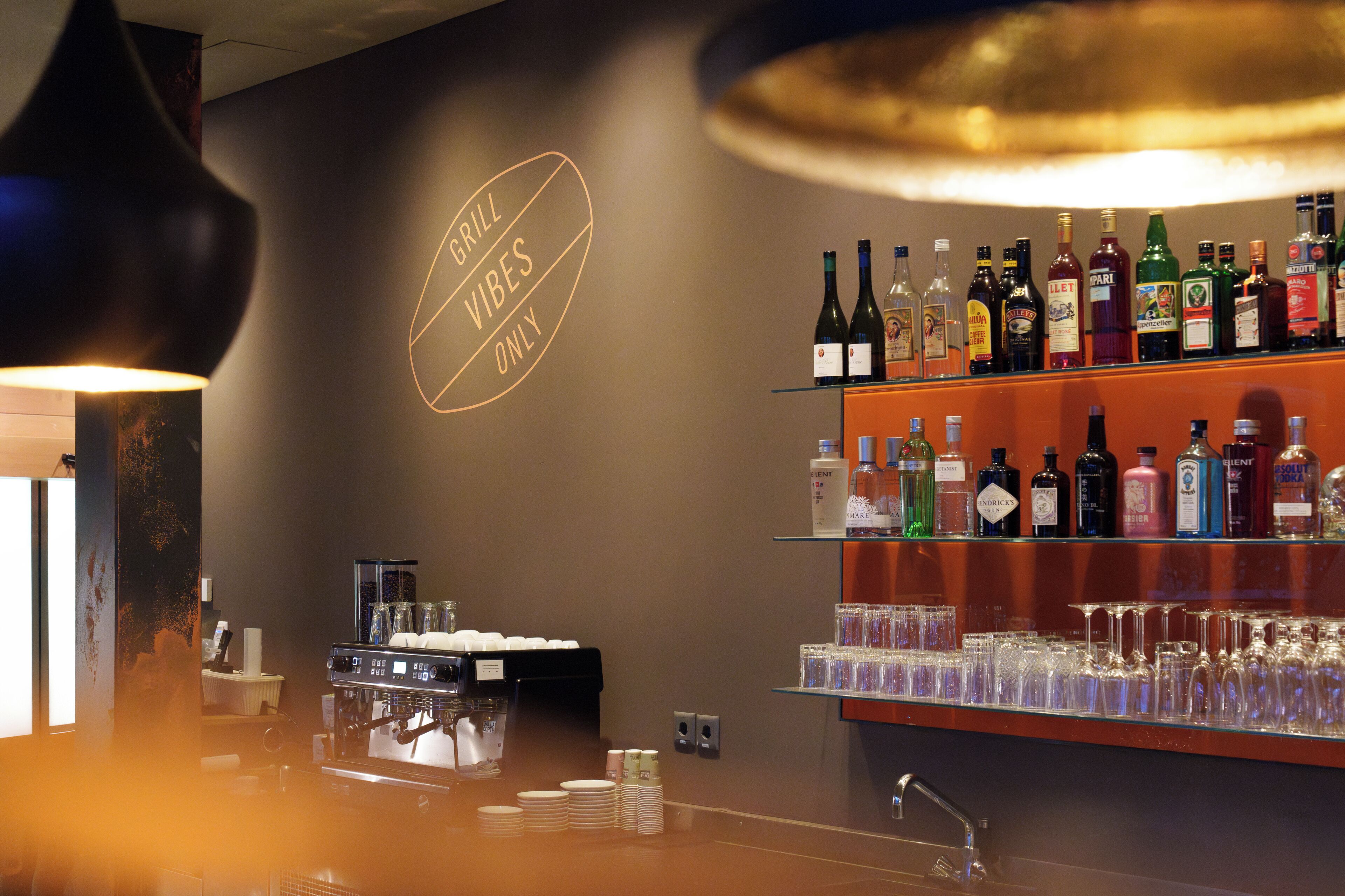 Uma área de bar com um letreiro escrito 'GRILL VIBES ONLY', prateleiras com diversas garrafas de bebidas e uma máquina de café.