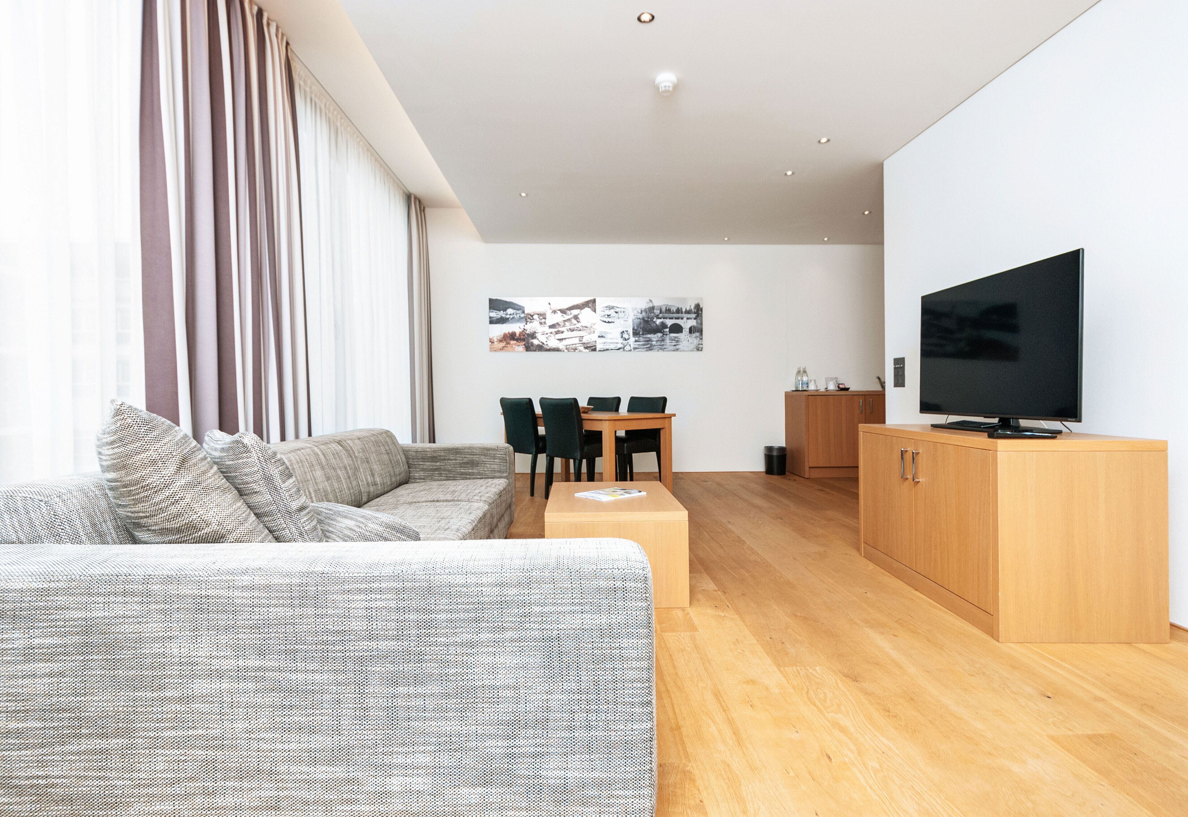 Uma sala de estar moderna com sofá, televisão, piso de madeira e área de jantar com cadeiras e mesa.
