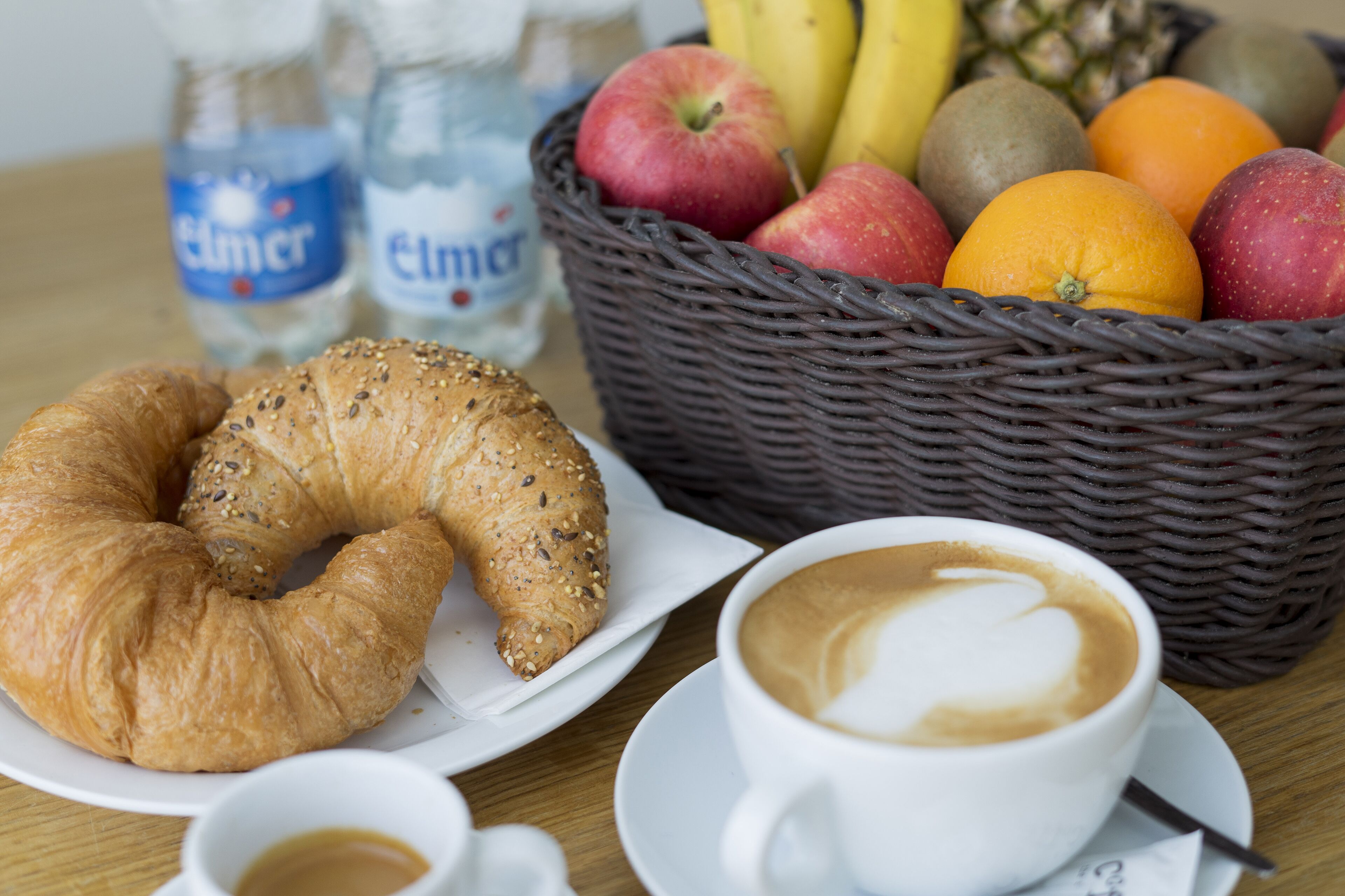 Una escena de desayuno con cruasanes, café, una cesta de frutas y botellas de agua.