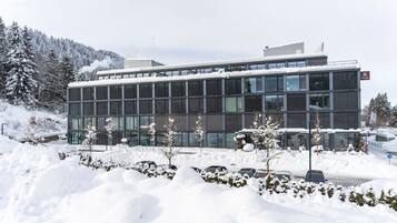 A modern building with a glass facade, surrounded by snow-covered trees and parked cars.