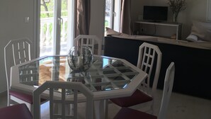 A dining area with a glass table, white chairs, and a glass bowl with candles. There are curtains on the windows, and a TV is visible in the background. - The Olive Grove House, in Kassiopi with all amenities in 5 min walking distance! (Κερκυρα)