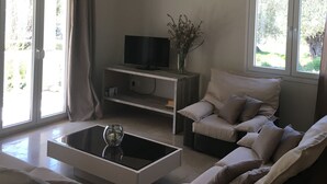 A living room with a sofa, a coffee table, a TV on a stand, and a chandelier. - The Olive Grove House, in Kassiopi with all amenities in 5 min walking distance! (Κερκυρα)