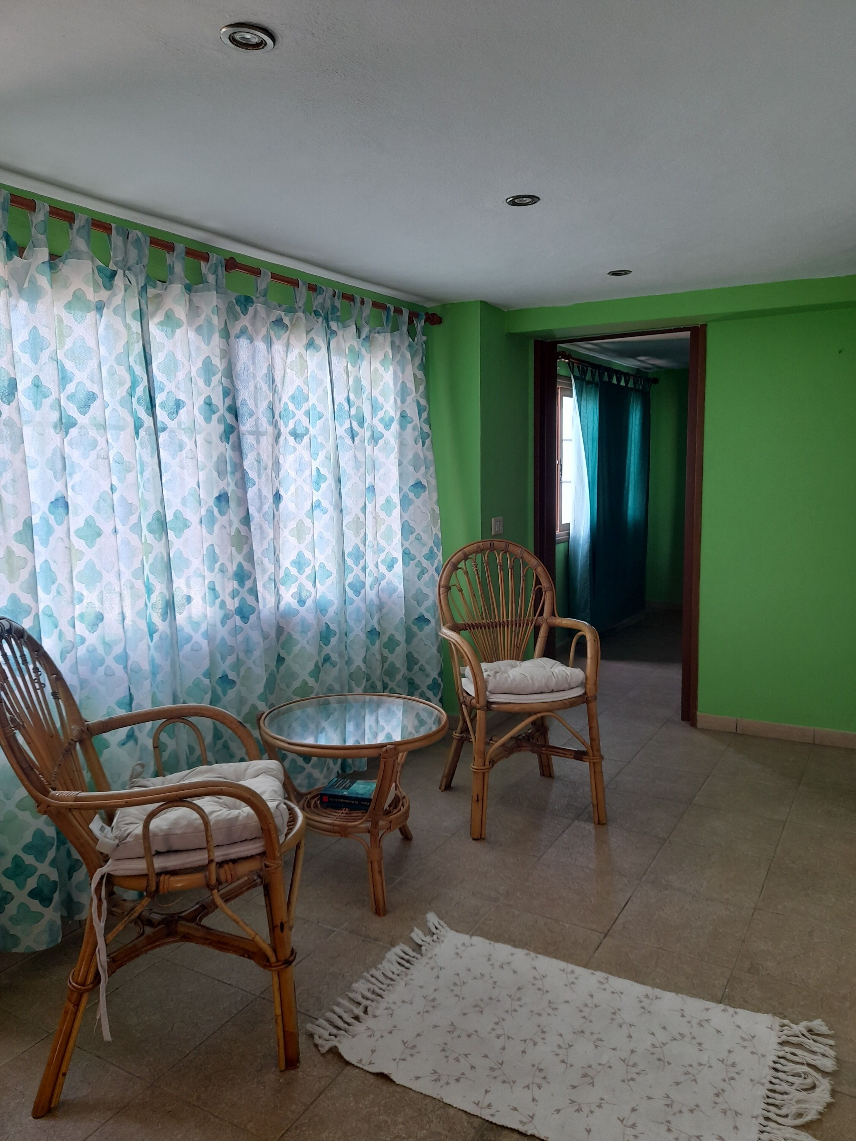 Une pièce comprenant deux fauteuils en osier, une table en verre et un tapis à motifs posé sur un sol carrelé.