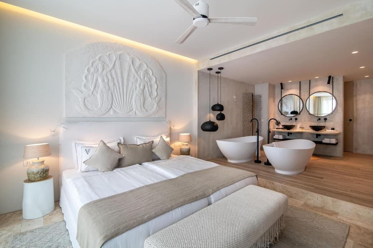 Ein modernes Schlafzimmer mit einem großen Bett, einem Deckenventilator, einer Wandskulptur, einer Nachttischlampe und einer geräumigen Badewanne im Badezimmer.