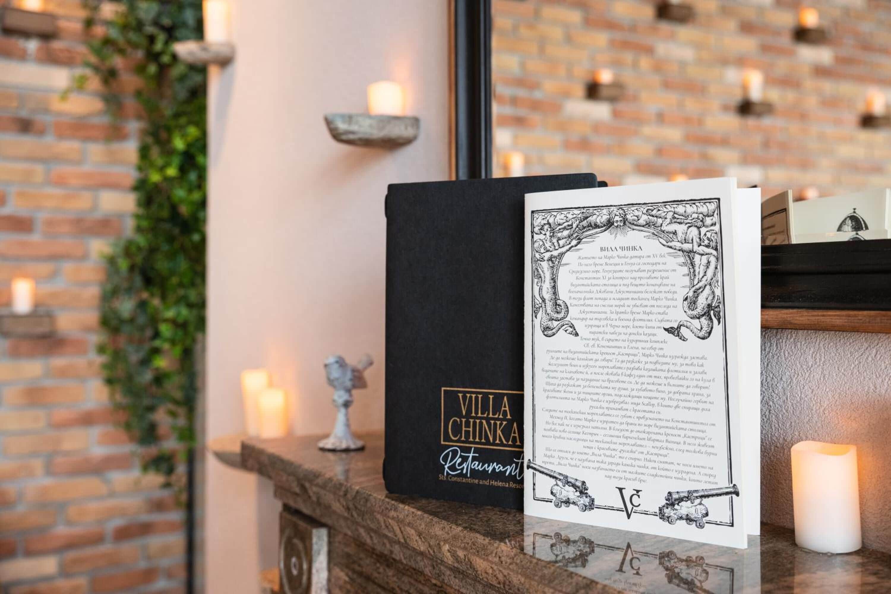 A black book titled 'Villa Chinka Restaurant' on a wooden surface with a candle and a small statue.