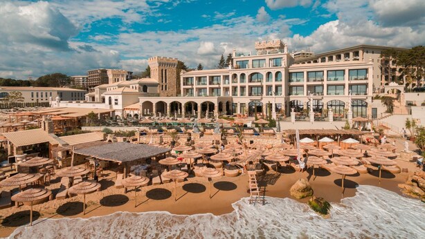 A beach resort with multiple sun umbrellas, a large building with arched windows, and a castle-like structure in the background. - Villa Chinka by Astor Garden Hotel (Varna)