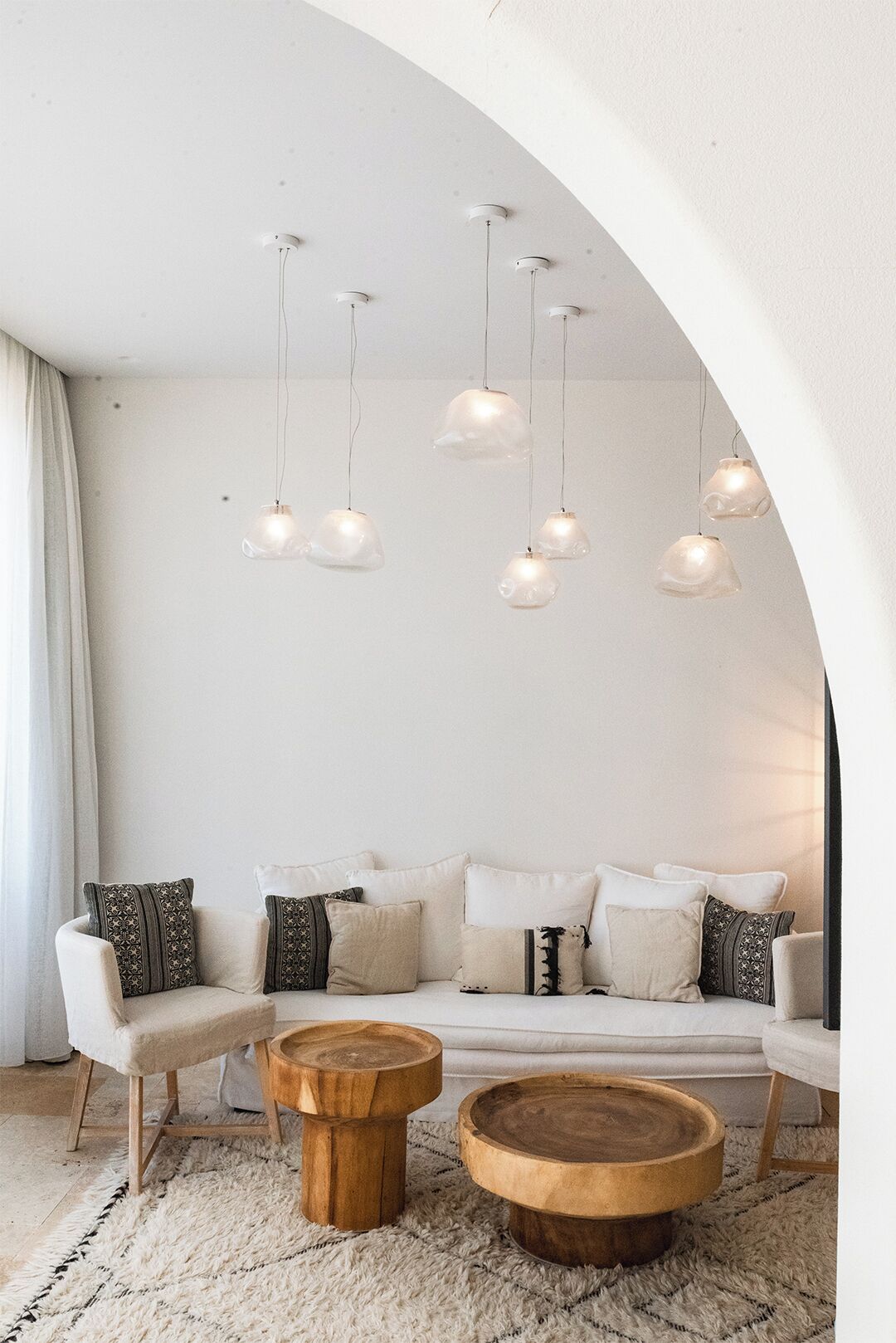 Un salon moderne avec un canapé blanc, des coussins à motifs, des tables d’appoint en bois et un design de plafond original agrémenté de luminaires suspendus.